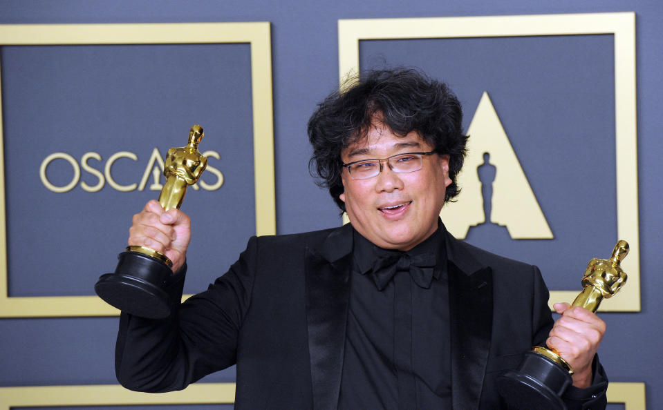 Individual in black suit holds two Oscars in front of event backdrop