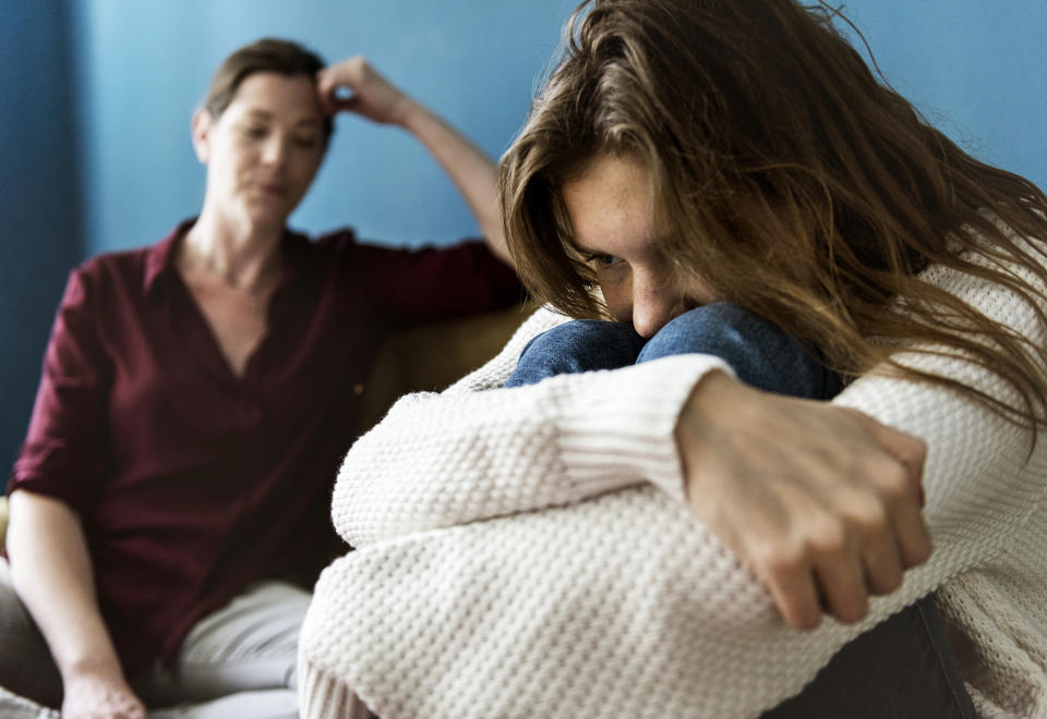 Mother and teenage daughter in candid moment; daughter, dressed in a sweater, appears distressed, hugging knees on couch