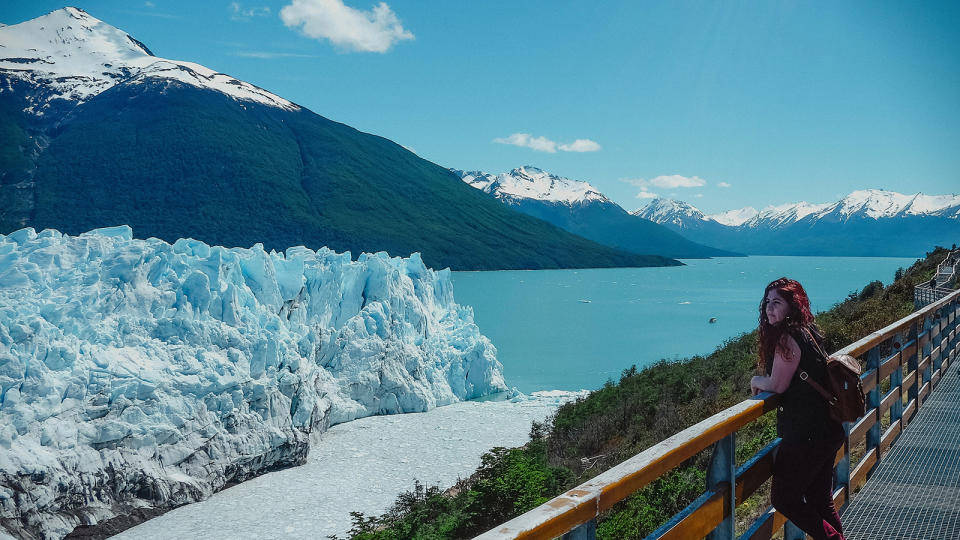 Woman standing on a bridge while looking at a glacier beside water and mountains