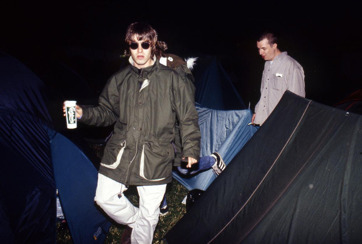 Liam Gallagher of Oasis backstage, holding a cup and wearing a parka, with brother Paul Gallagher, Glastonbury Festival, 1995.