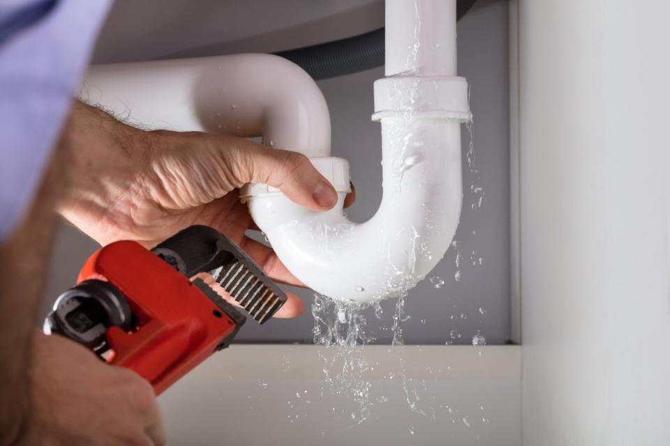 Person holding a red wrench, fixing a leaking PVC pipe under a sink.