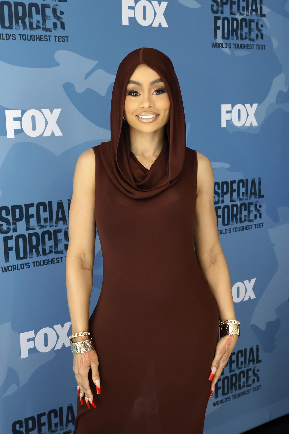 Person in a brown dress with a draped neckline and matching headscarf posing on a promotional backdrop