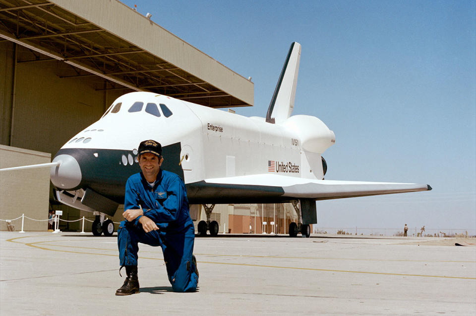 a man in a blue flight suit is kneeling in front of a white space shuttle on a runway