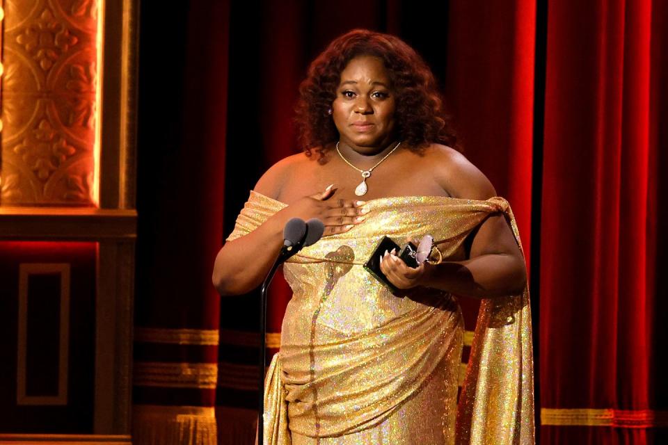 alex newell wears a gold dress while holding a tony award and speaking into a microphone on a stage, with a red curtain behind them
