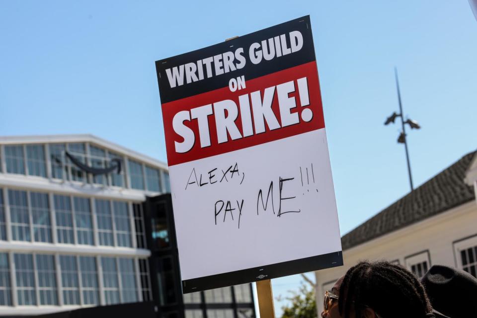 A protester holds a sign that says, "Writers Guild on Strike." Below it says "Alexa, Pay Me "