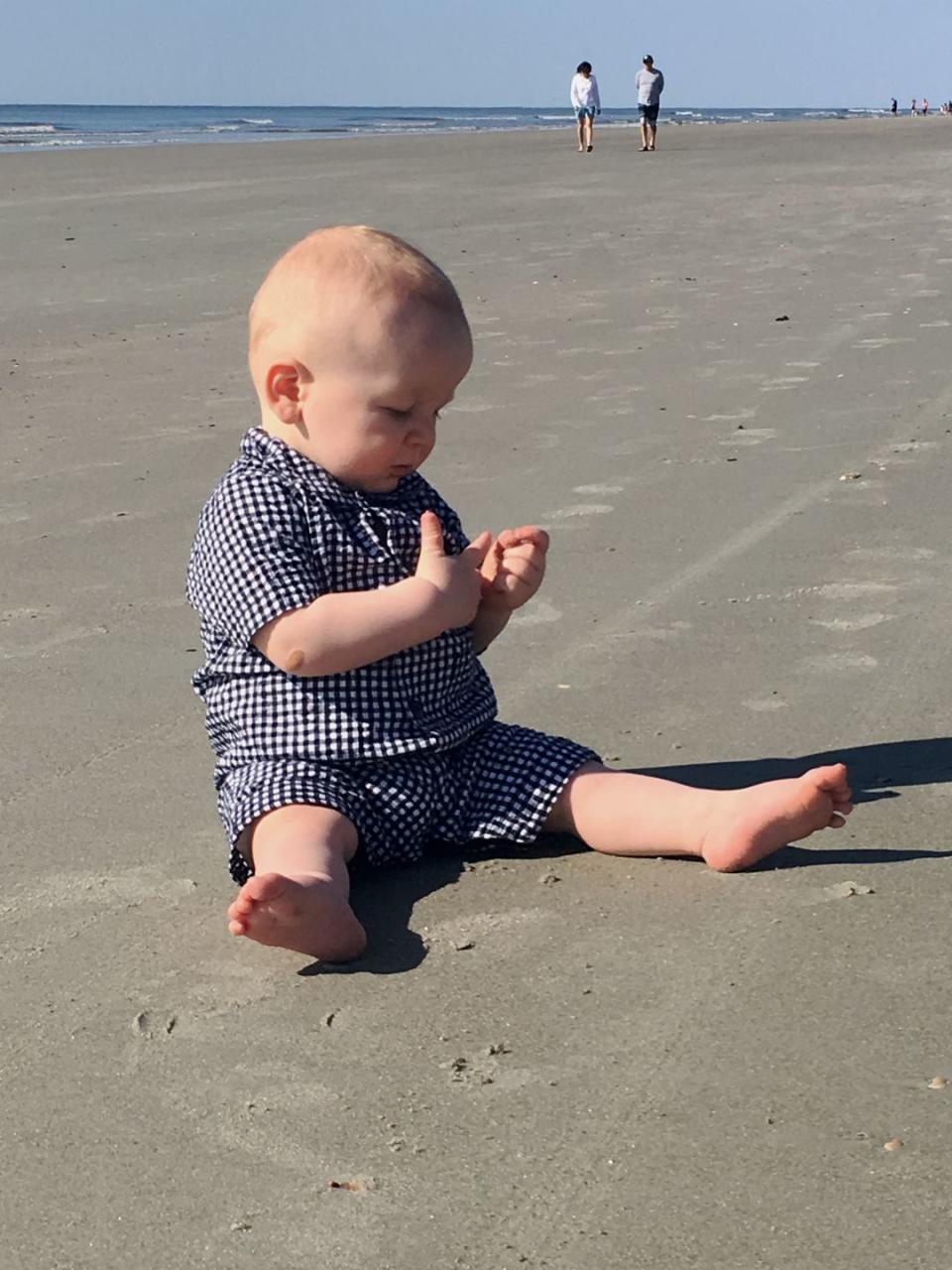 baby boy in blue and white plaid on the beach with people in the background