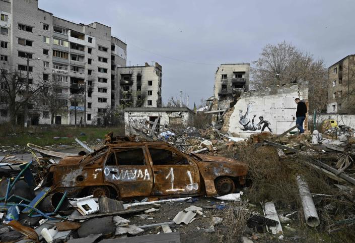 A destroyed car and other rubble is seen in the foreground as burned out buildings, including a wall with a mural, are seen behind.