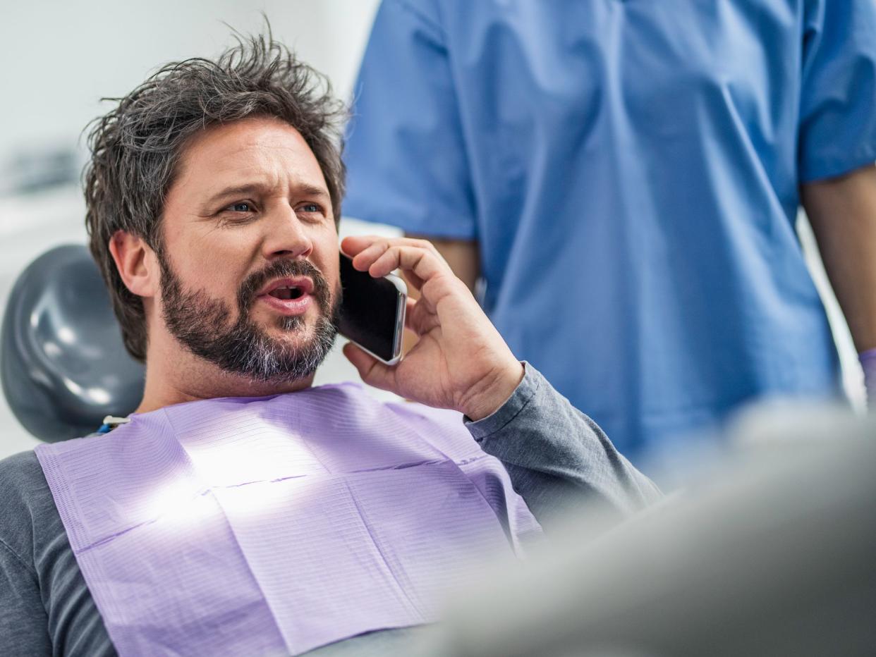 Men sitting in dentist’s chair and talking on a smart phone, dentist assistant standing behind him.