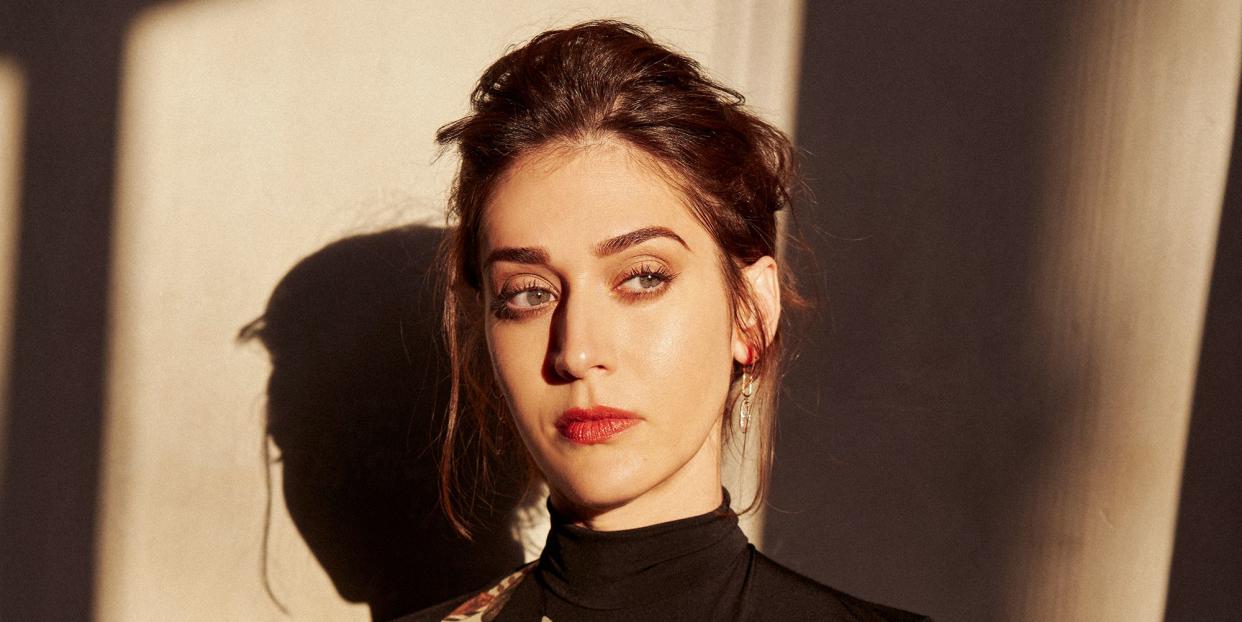 actor lizzy caplan shown in a dramatic portrait with her hair up, eyebrow arched, in a black and floral bodysuit tucked into jeans