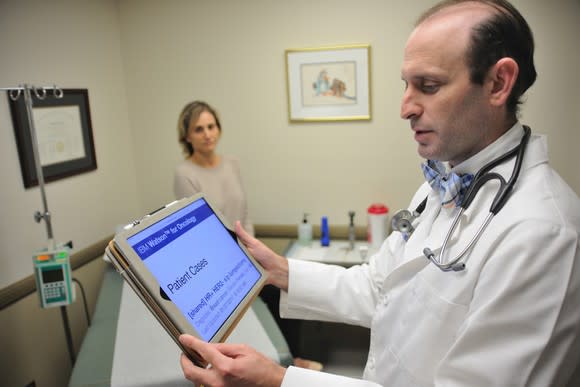 A doctor using a tablet computer, with a patient in the background