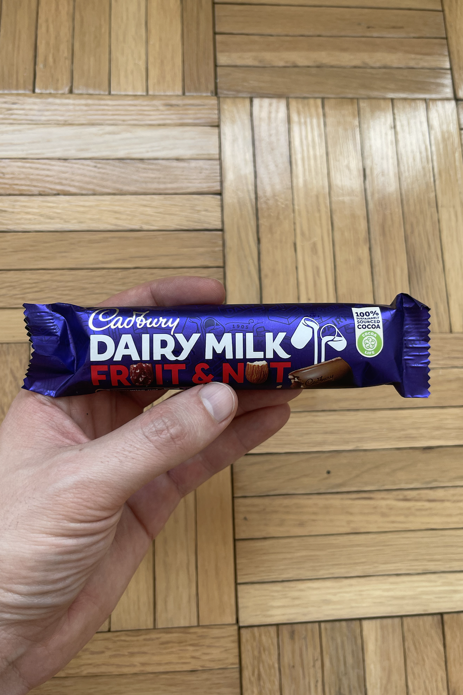 A hand holding a Cadbury Dairy Milk Fruit & Nut chocolate bar over a wooden floor