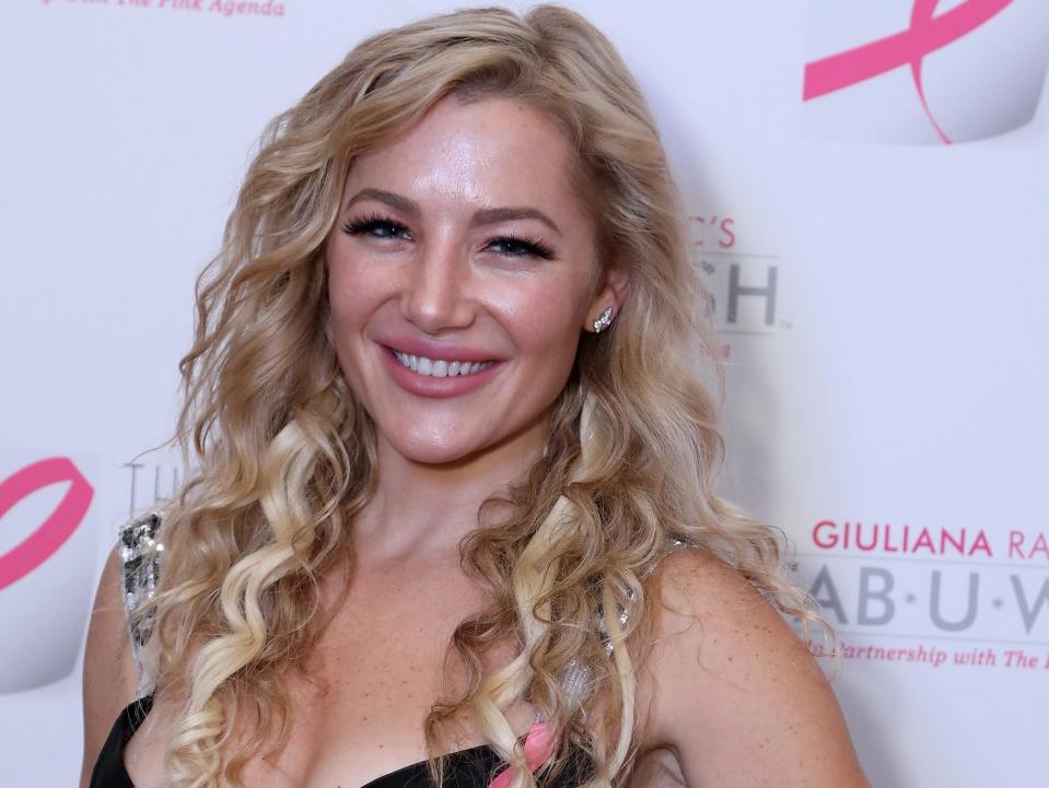 Hayley Paige smiles in a black dress in front of a white and pink backdrop.