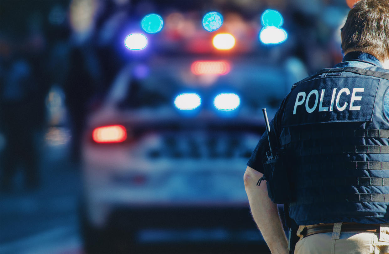 A police officer's back is shown as he looks toward a patrol car with flashing lights.