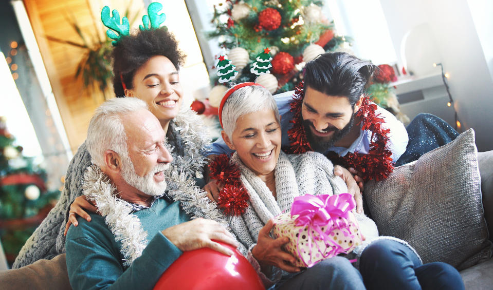 Closeup front view a senior couple sharing Christmas presents with their grandson and his fiancee. They are comfortably seated on a living room sofa, Christmas tree in background.