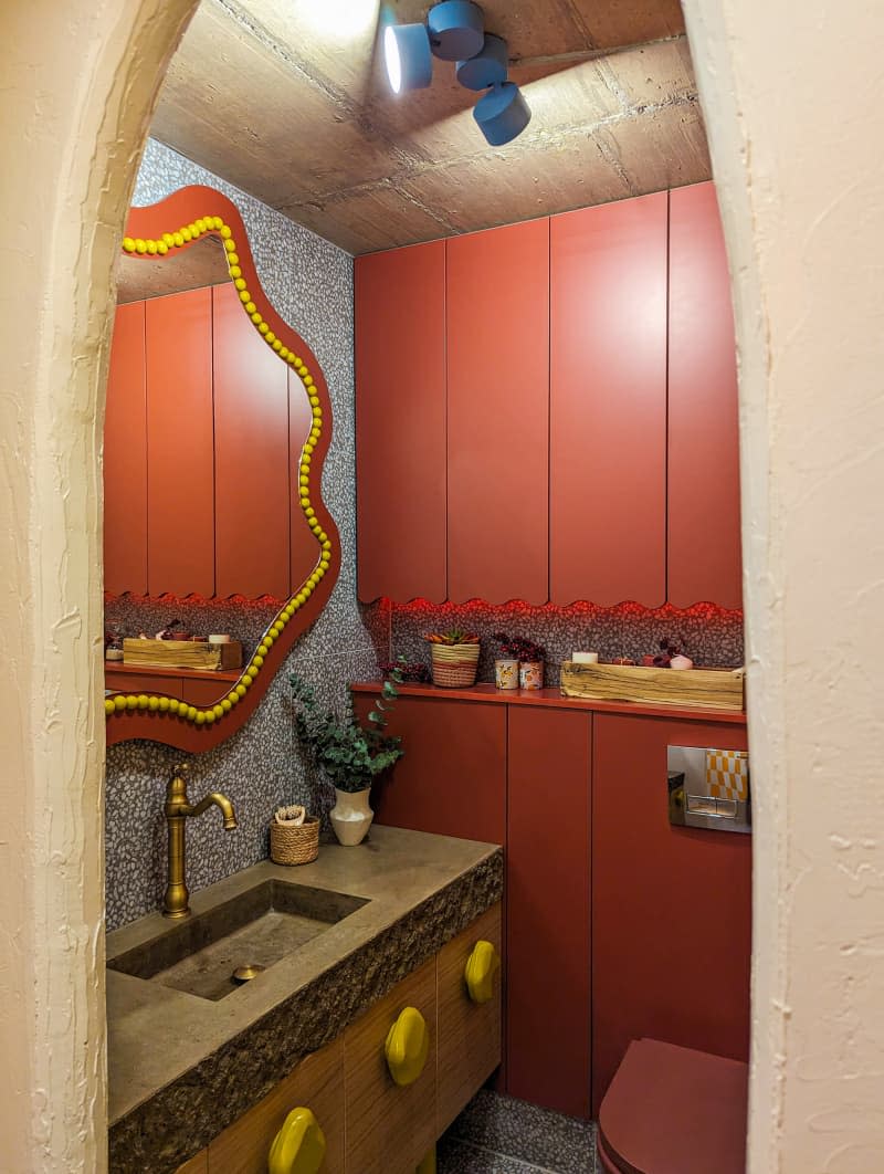 Bathroom with wavy orange and yellow mirror, wood cabinets with stone slab counter and yellow irregular shaped knobs, and lots of orange cabinets