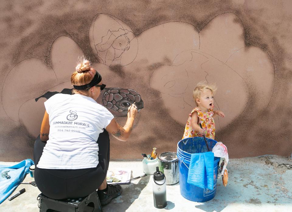 Summer DeSalvo paints the bottom of a mural on Fort Myers Beach while her daughter, Nova, 1, plays with her paintbrushes on Monday, July 24, 2023. The mural is on a three-story elevator shaft left standing after Hurricane Ian in Times Square. Summer painted a sunset scene of the pier at the top of the mural last Friday. She plans to be done with this wall and the wall adjacent to it by the end of the week, and another artist, Lacy McClary, is painting the other two sides.