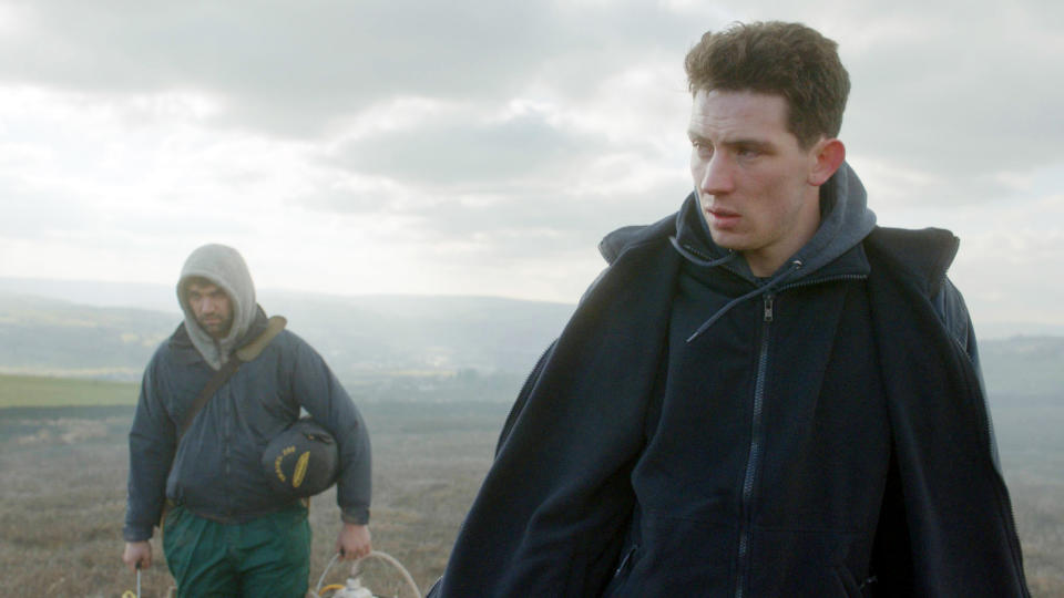 Josh O'Connor and Alec Secareanu walking in a moorland setting, with O'Connor wearing a jacket and hoodie, and Secareanu carrying a bag