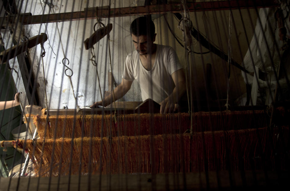 In this Saturday, July 6, 2019 photo, Palestinian worker weaves carpets on a traditional wooden loom at a carpets factory in Gaza City. Talk about old Gaza, and what pops up are images of clay pottery, colorful glassware, bamboo furniture and ancient frame looms weaving bright rugs and mats. As such professions could be dying worldwide, the pace of their declining is too fast in Gaza that out of its some 500 looms, only one is still functioning. (AP Photo/Khalil Hamra)