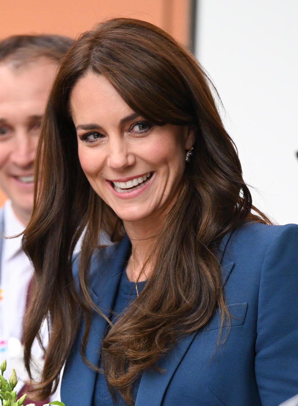 Kate Middleton smiling and wearing a blue blazer. Another person is partially visible in the background, smiling