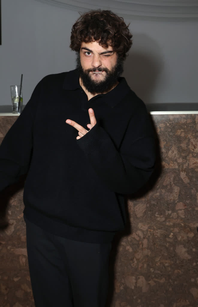 Person posing with a peace sign, wearing a black sweater and sporting facial hair