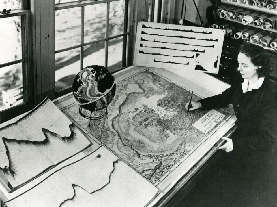 Geologist Marie Tharp sits at a large drafting table holding a pen and working on a map of the Atlantic Ocean floor.