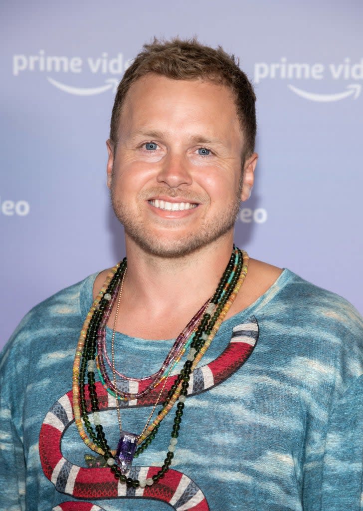 Spencer Pratt posing at an event, wearing a blue patterned T-shirt and multiple beaded necklaces