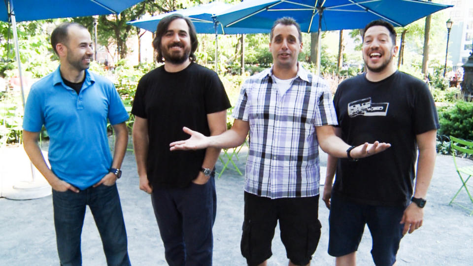 Four men standing under an umbrella, smiling, casual attire