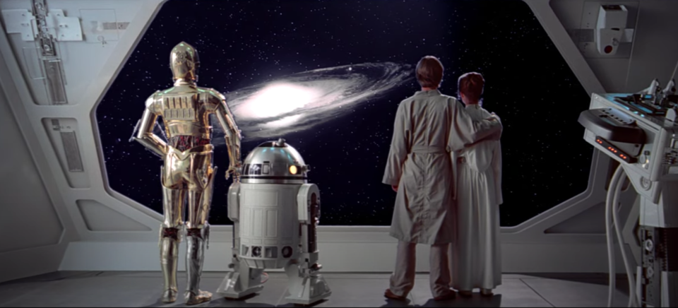 Two droids, a man, and a woman, stand together, looking out the back of a space ship