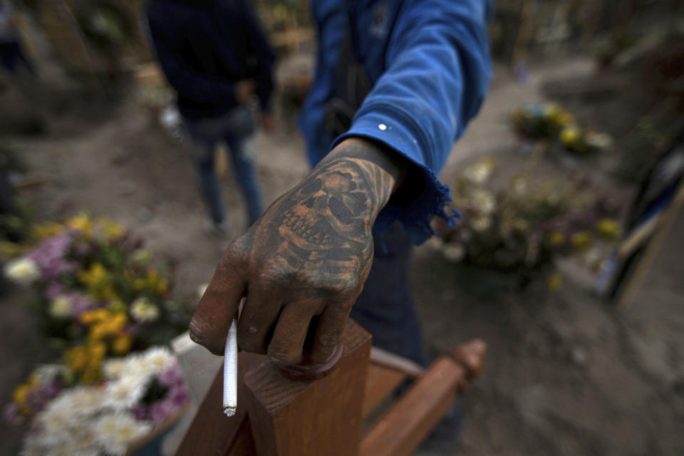 FILE - In this March 17, 2021 file photo, a cemetery worker, donning a skull tattoo on his hand, holds a cigarette during the burial service of a person who died from COVID-19, in the Chalco cemetery on the outskirts of Mexico City. As Mexico approaches 200,000 in officially test-confirmed deaths from COVID-19, the real death toll is probably higher due to the country’s extremely low rate of testing. (AP Photo/Fernando Llano, File)