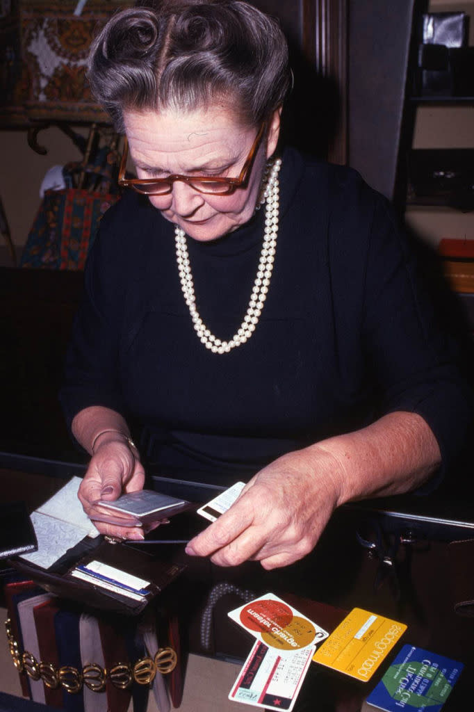 A woman with glasses and a pearl necklace examines multiple credit cards and IDs atop a table