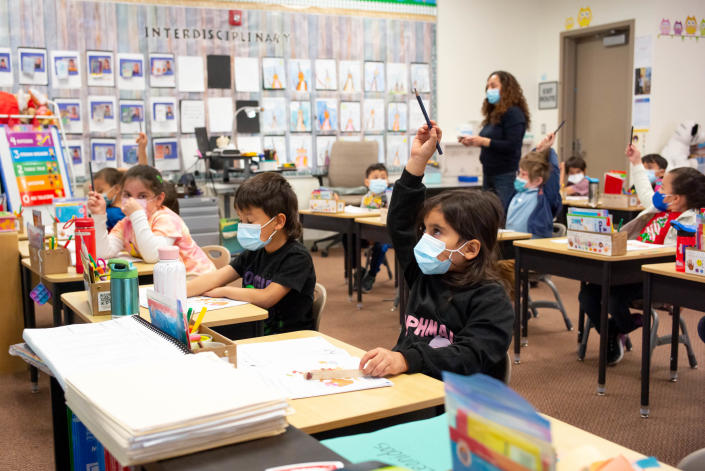 Young students are seated at desks in a classroom with some raising pencils above their heads. A teacher stands near the door. All wear face masks.