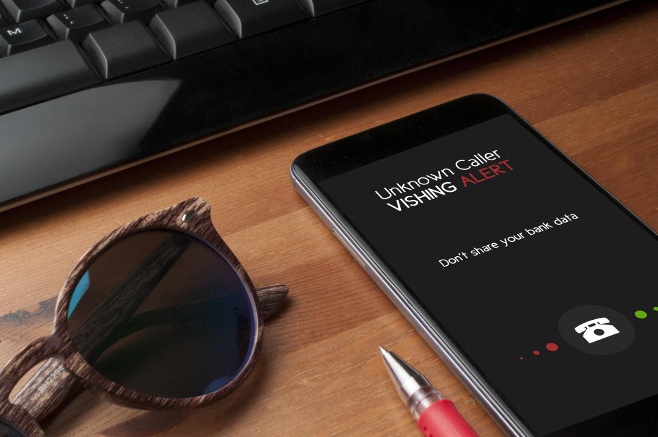 Vishing (voice phishing) concept, a smartphone on a table next to a computer keyboard and a pair of sunglasses show an unknower caller call with vishing alert and a reminder to not share personal bank data