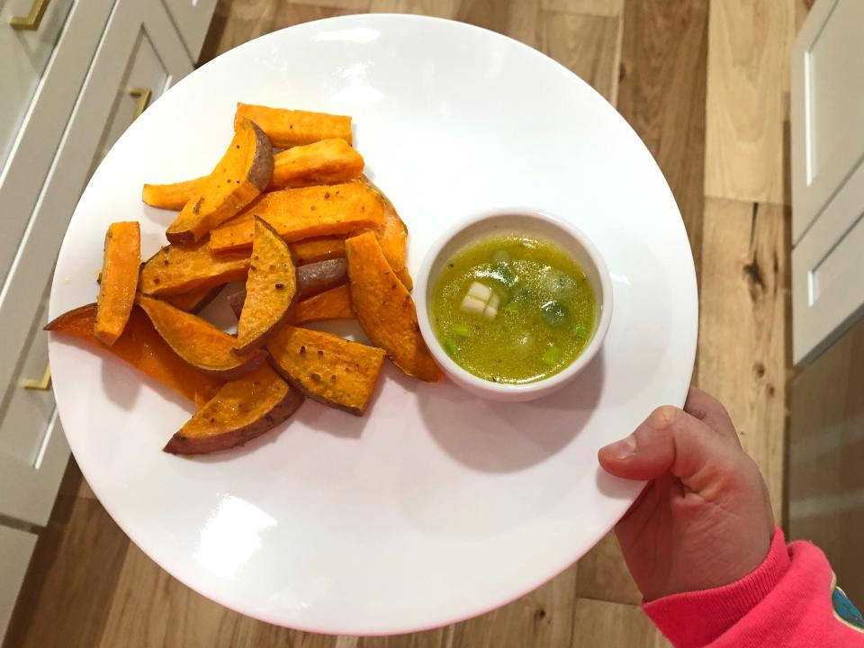A hand holding a white plate with sliced sweet-potato wedges. On the plate is a ramekin with Ina Garten's vinaigrette mixed with scallions.