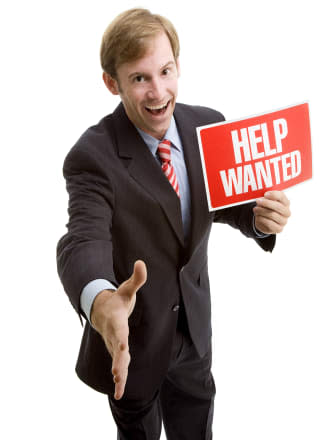 A handsome young businessman stands holding a Help Wanted sign with his hand outstretched for a handshake.