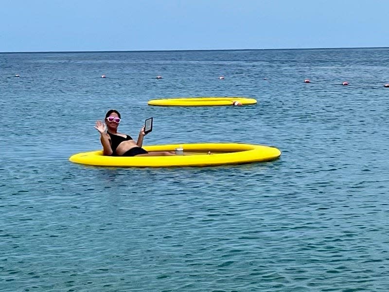 Terri Peters is reading her Kindle while wearing a bathing suit and sunglasses and swimming in the water in a yellow raft.