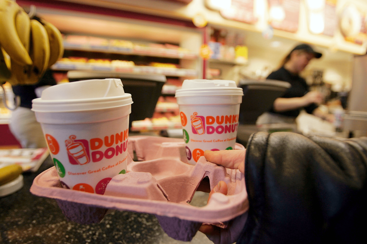 Focus on a tray of two travel cups of coffee in a Dunkin' Donuts, Cambridge, Massachusetts, a customer carrying it, cashier and rows of donuts blurred in the background