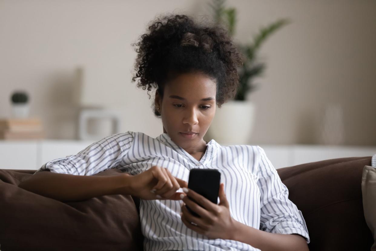 Focused African American young woman using smartphone, sitting on couch, confident young female looking at phone screen, typing writing message in social network, browsing apps, chatting online