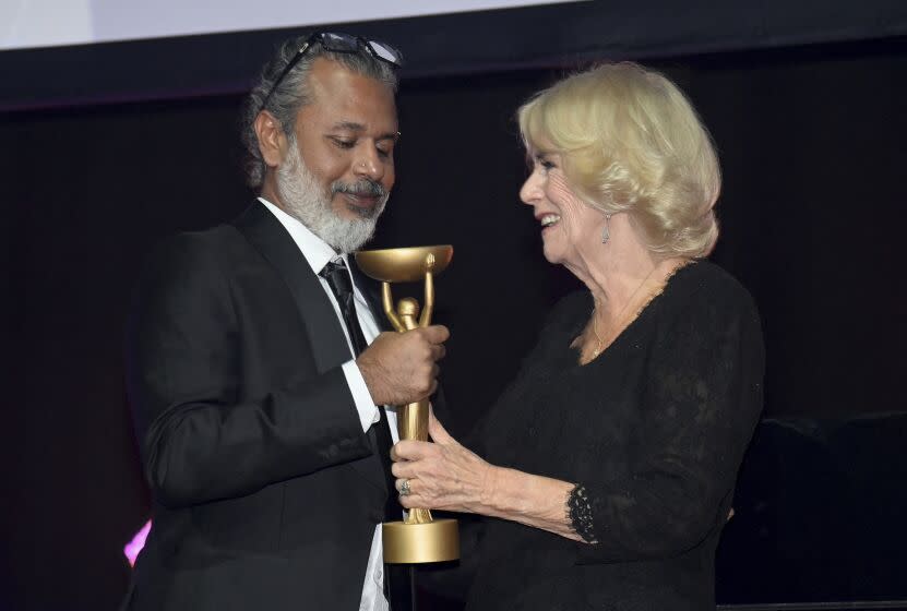 A man with gray hair and a beard and blond woman, both in formalwear, gripping a gold trophy