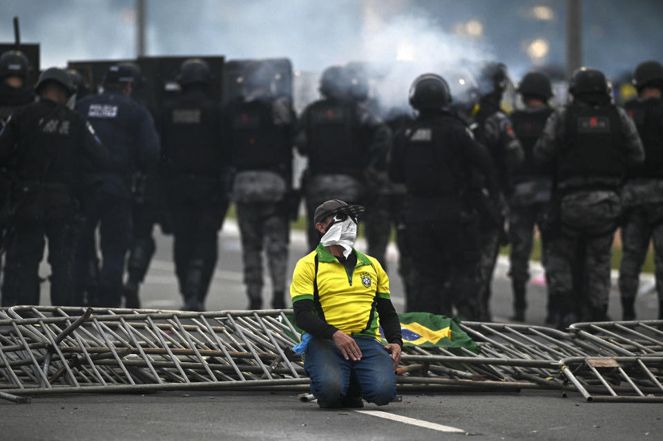 A person wearing a green and yellow shirt and white bandana over their face kneels next to fallen crowd-control fencing behind a few dozen police in riot gear.