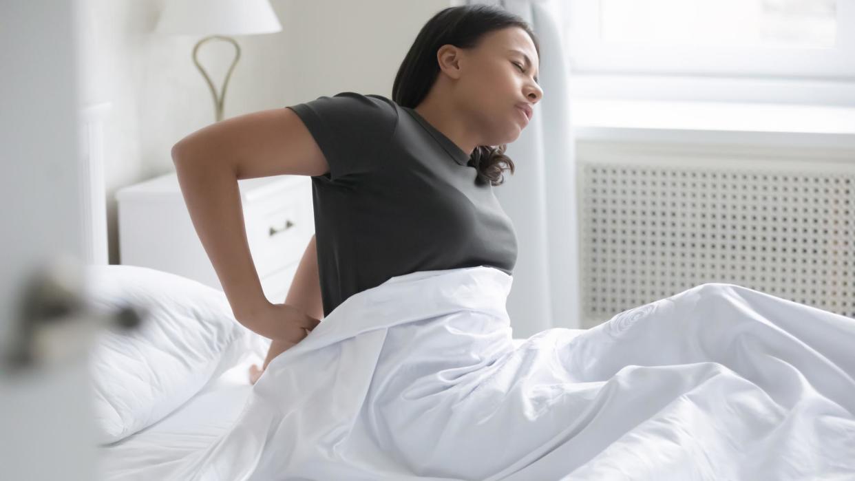  A woman in a brown t-shirt massages her painful lower back because she's sleeping on a memory foam mattress that's too soft for her and she should switch to a hybrid bed. 