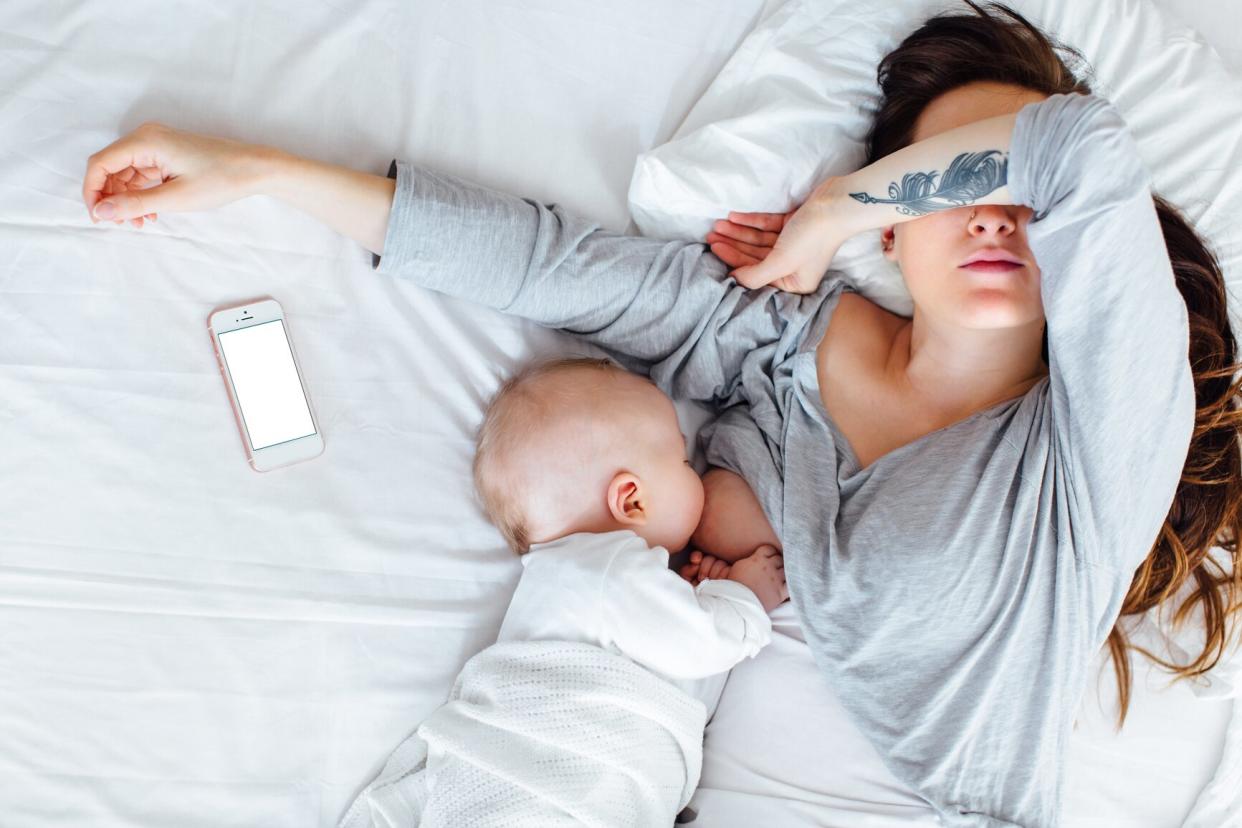 A Tired Mother Covering Her Eyes As She Breastfeeds Her Baby Boy Lying Down On A Bed