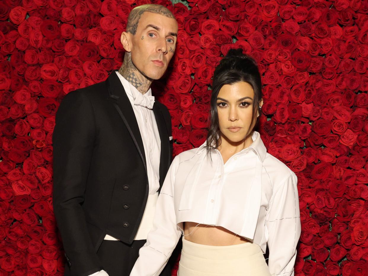 Kourtney Kardashian, who is wearing cream-colored pants and a white crop top, and Travis Barker, who is wearing a black suit and a white bowtie, posing together in front of a wall of roses.