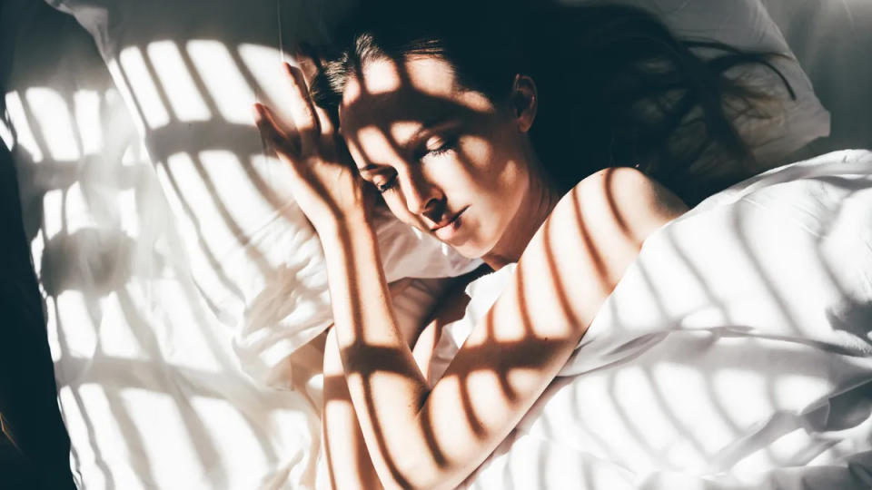 A woman with long dark hair sleeps under a white duvet with her bare arms on show