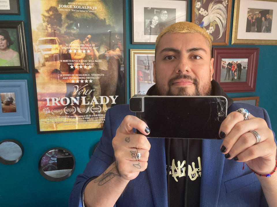 Jorge Xolalpa, a 33-year-old award-winning Mexican-born filmmaker holds his iPhone with his film "Your Iron Lady," posters and framed magazine articles on wall at his home on Monday, Oct. 10, 2022 in San Pedro, Calif. Xolalpa is mired in a years-long battle over whether he can keep working legally in the United States. He is among hundreds of thousands of people waiting to learn if the program known as Deferred Action for Childhood Arrivals will be allowed to continue. (AP Photo/Eugene Garcia)