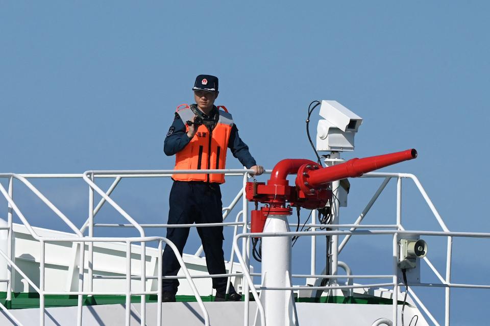 A photo of a man in a hat and life vest standing with his left hand on a railing and his right hand holding a camcorder.