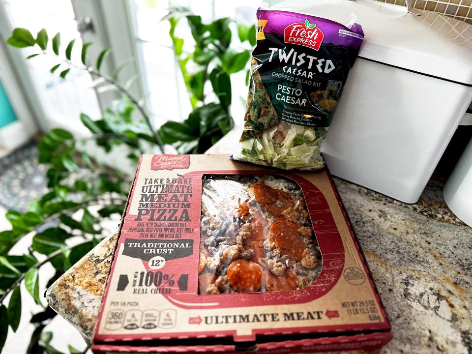 A cardboard box with red detailing containing a meat pizza next to a black and purple bag of chopped salad on a stone countertop