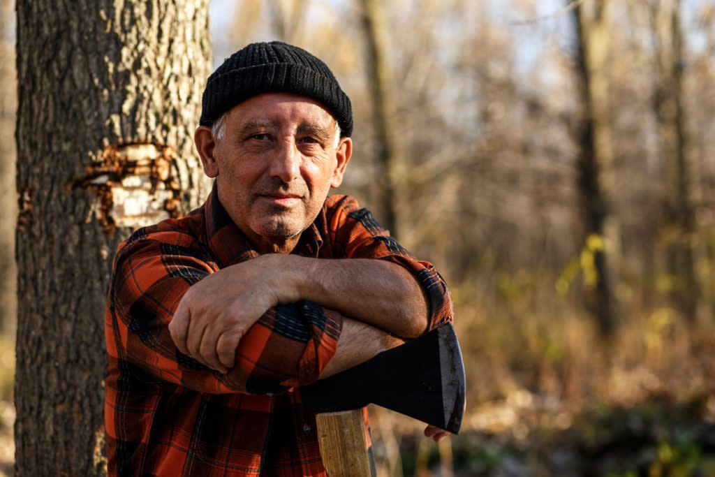 A senior citizen poses for a portrait with an axe and tree behind him.