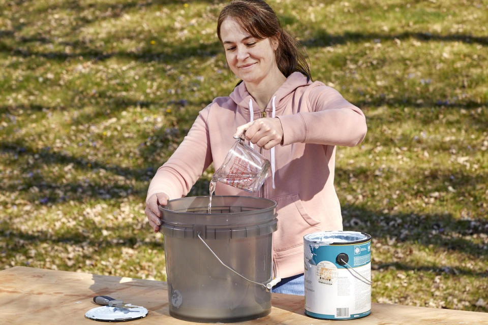 Woman pours a cup of water into a large bucket, a can of white paint next to the bucket.