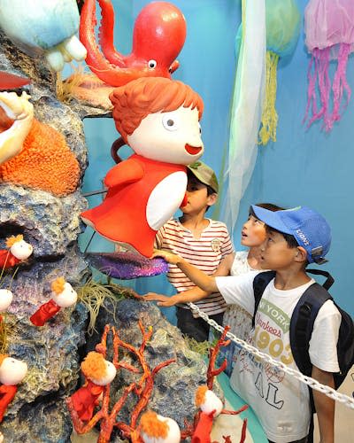 Three children stare up at a large, very colorful structure that looks like a coral reef with clay characters on it.