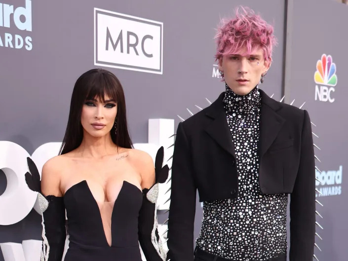megan fox in a low cut dress with a thigh slit and flowers on the sleeves with machine gun kelly, wearing a sparkling shirt and a cropped blazer with spikes on the shoulders and arms on the billboard music awards red carpet